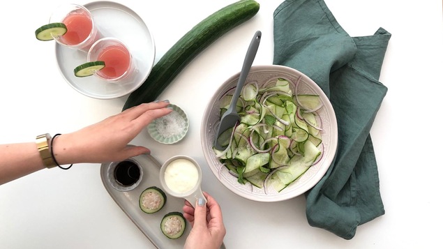 Vue en plongée de verres de jus de concombre et de pastèque, des "sushis" de concombres et une salade de rubans de concombre avec une main qui tend de la vinaigrette.