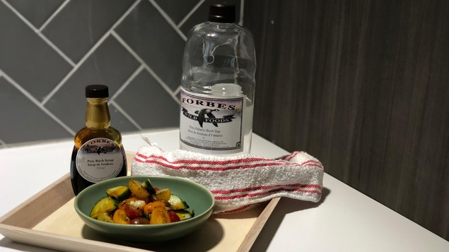 Un plateau avec une bouteille de sirop de bouleau, une bouteille de sève de bouleau et une salade de tomates et de concombres.
