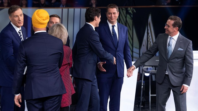 Cinq politiciens et une politicienne se serrent la main sur un plateau de télévision.