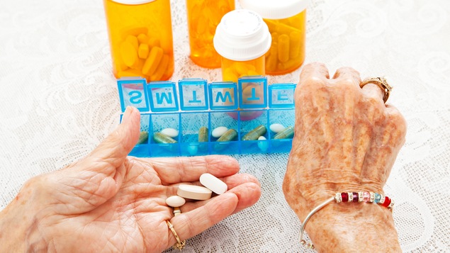 Les mains d'une vieille dames tiennent des médicaments variés. En arrière-plan, on voit un pilulier et des contenants de médicaments.