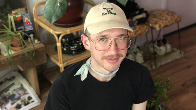 Alasdair Rees assis dans son salon. Il porte un chapeau et on voit des plantes derrière lui.
