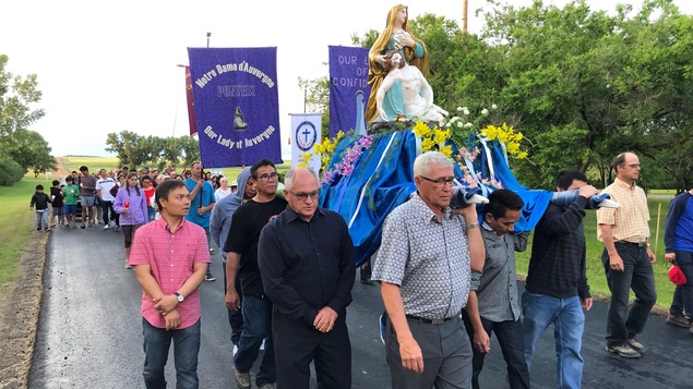 Des hommes portent une base recouverte de tissu bleu, sur laquelle est installée une statue de la Vierge Marie. Ils sont suivis par un groupe de fidèles.