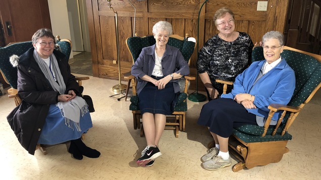 Quatre sœurs religieuses ont de grands sourires. Trois sont assises sur des chaises à bascule, la quatrième est agenouillée sur le plancher.