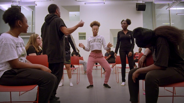 Sous les yeux d'un chorégraphe et d'autres jeunes filles assises, une jeune fille fait un mouvement de danse Sous les yeux d'un chorégraphe et d'autres jeunes filles assises, une jeune fille fait un mouvement de danse