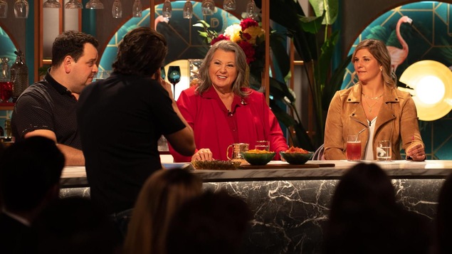 L'animateur, de dos, s'adresse à Ginette Reno, souriante, qui est de l'autre côté d'un comptoir. L'animateur, de dos, s'adresse à Ginette Reno, souriante, qui est de l'autre côté d'un comptoir.
