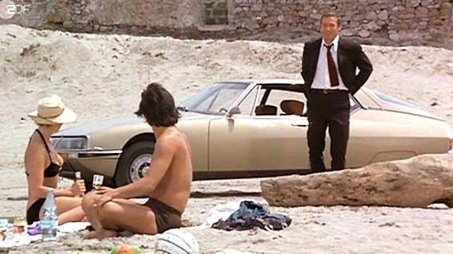 Un couple assis sur la plage en maillots de bain regarde un autre homme en costume, devant sa voiture Un couple assis sur la plage en maillots de bain regarde un autre homme en costume, devant sa voiture