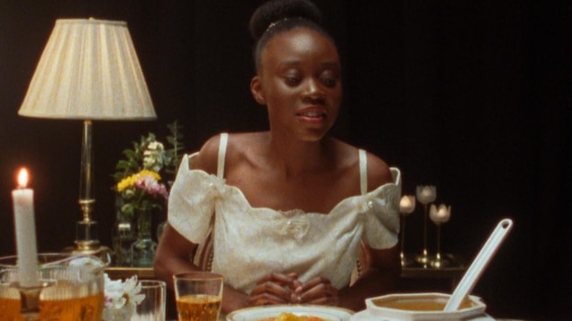 Une jeune femme (Schelby Jean-Baptiste) assise devant un festin sur une table.