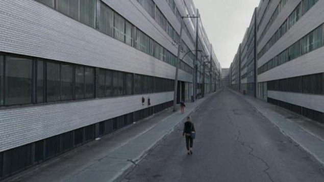 A woman from behind is walking between two rows of gray buildings.