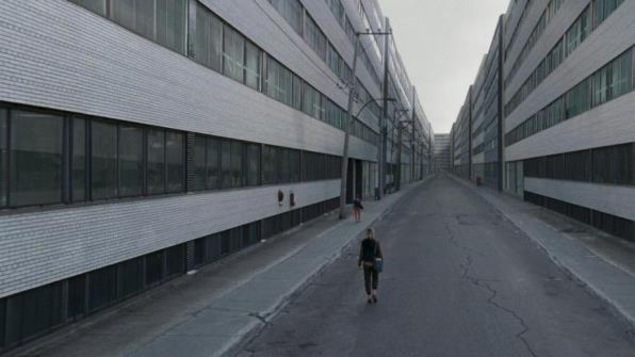 Une femme de dos marche entre deux rangées d'immeubles gris.