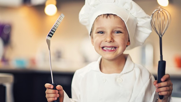 Il porte une toque de chef. Il tient dans ses mains une spatule et un fouet. Il porte une toque de chef. Il tient dans ses mains une spatule et un fouet.