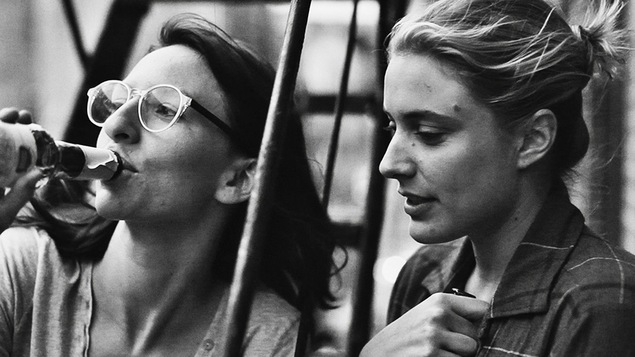 En noir et blanc, deux jeunes filles (dont Greta Gerwig) boivent une bière sur des escaliers extérieurs. En noir et blanc, deux jeunes filles (dont Greta Gerwig) boivent une bière sur des escaliers extérieurs.
