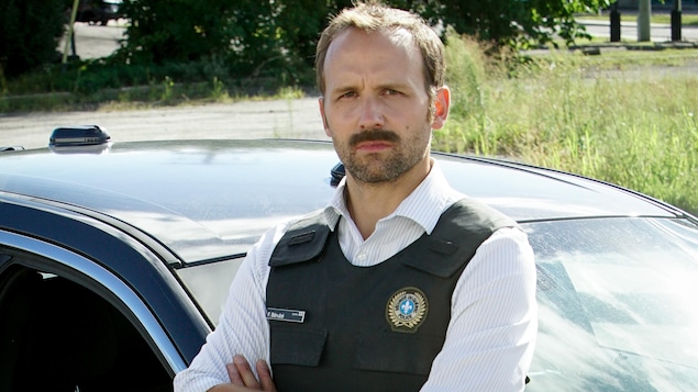 Frédérik « Fred » Bérubé (Émile Proulx-Cloutier) affiche un air sérieux. Il porte une veste sur laquelle on voit un écusson de policier.

