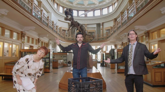 Chantal Lamarre, MC Gilles et Jean-René Dufort répondent à des questions dans un musée. À l'arrière, il y a un squelette de dinosaure.