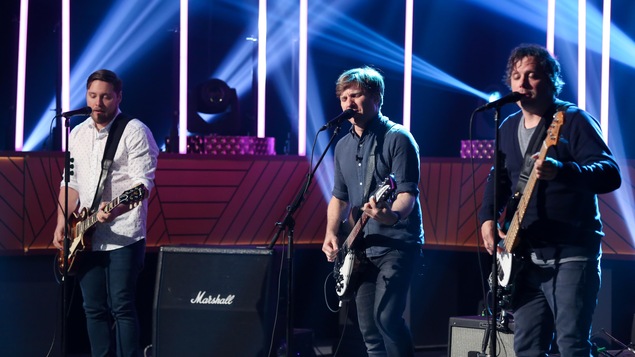 Trois membres du groupe au micro avec leurs guitares et basse.