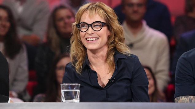 Elle sourit. Ses cheveux sont blonds-roux et elle porte des lunettes à montures noires. Elle porte un chemisier noir.