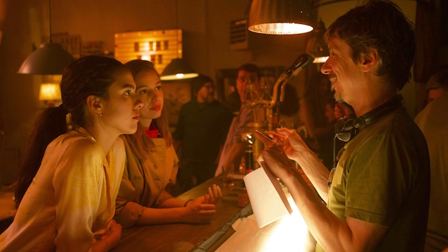 Dans un bar sous une lumière rouge, un homme derrière le bar portant des écouteurs autour du cou (Philippe Falardeau) parle à deux jeunes femmes accoudées sur le bar. Dans un bar sous une lumière rouge, un homme derrière le bar portant des écouteurs autour du cou (Philippe Falardeau) parle à deux jeunes femmes accoudées sur le bar.