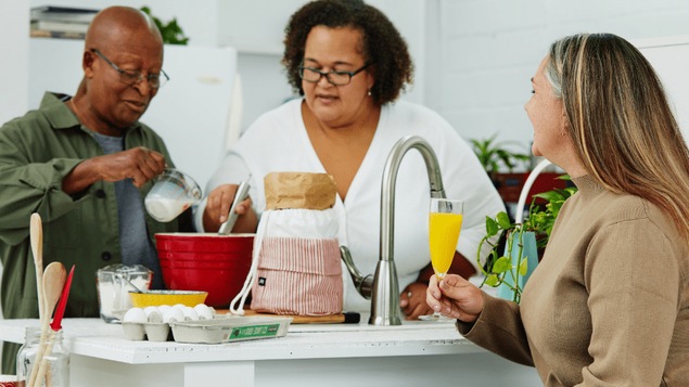 Trois personnes sont dans une cuisine. L'une est assise avec un verre à la main et les deux autres cuisinent derrière un comptoir. Trois personnes sont dans une cuisine. L'une est assise avec un verre à la main et les deux autres cuisinent derrière un comptoir.