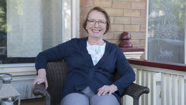 Le personnage de Rollande Mathieu, interprété par Marie-Ginette Guay, est assis sur une chaise devant sa maison et sourit à la caméra.