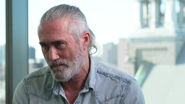 Roy Dupuis est assis dos à une fenêtre qui donne sur la ville de Montréal.