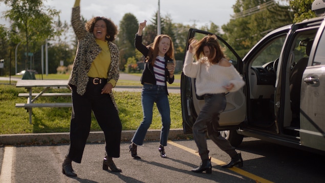 Trois femmes dans un stationnement dansent de joie un poing dans les airs.