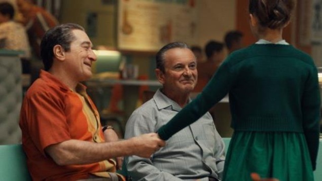 Un homme en chemise orange (Robert DeNiro) assis à côté d'un autre souriant (Joe Pesci), tient la main d'une petite fille de dos Un homme en chemise orange (Robert DeNiro) assis à côté d'un autre souriant (Joe Pesci), tient la main d'une petite fille de dos