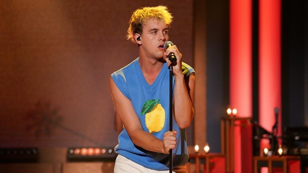 Un homme porte un chandail bleu qui affiche l'image d'un citron. Il chante dans un micro.