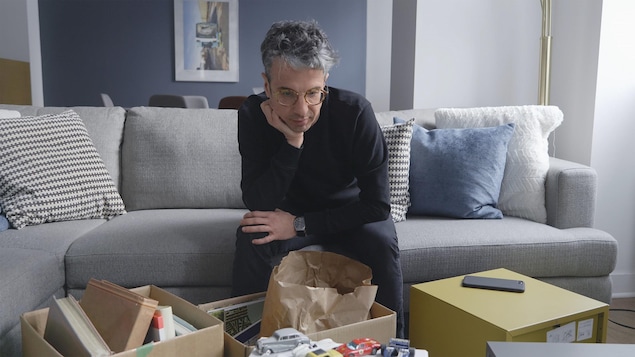 Matthieu Dugal est assis sur un canapé, l'air pensif, avec des cartons remplis d'objets devant lui.
