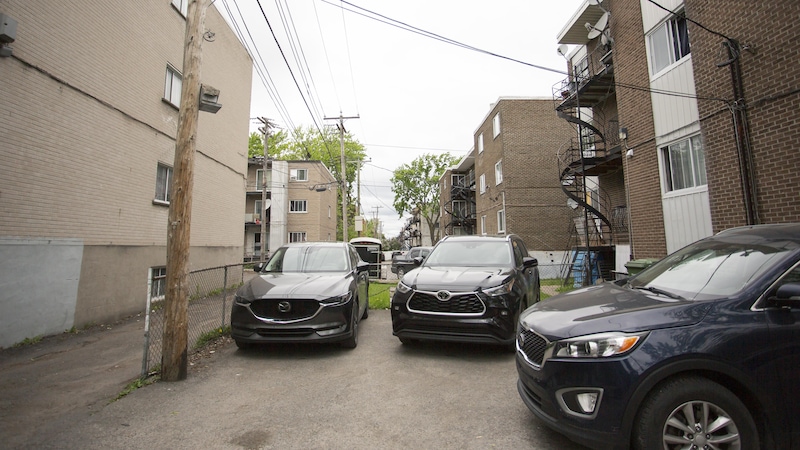 Des voitures sont stationnées dans un espace asphalté, derrière des immeubles à logements.