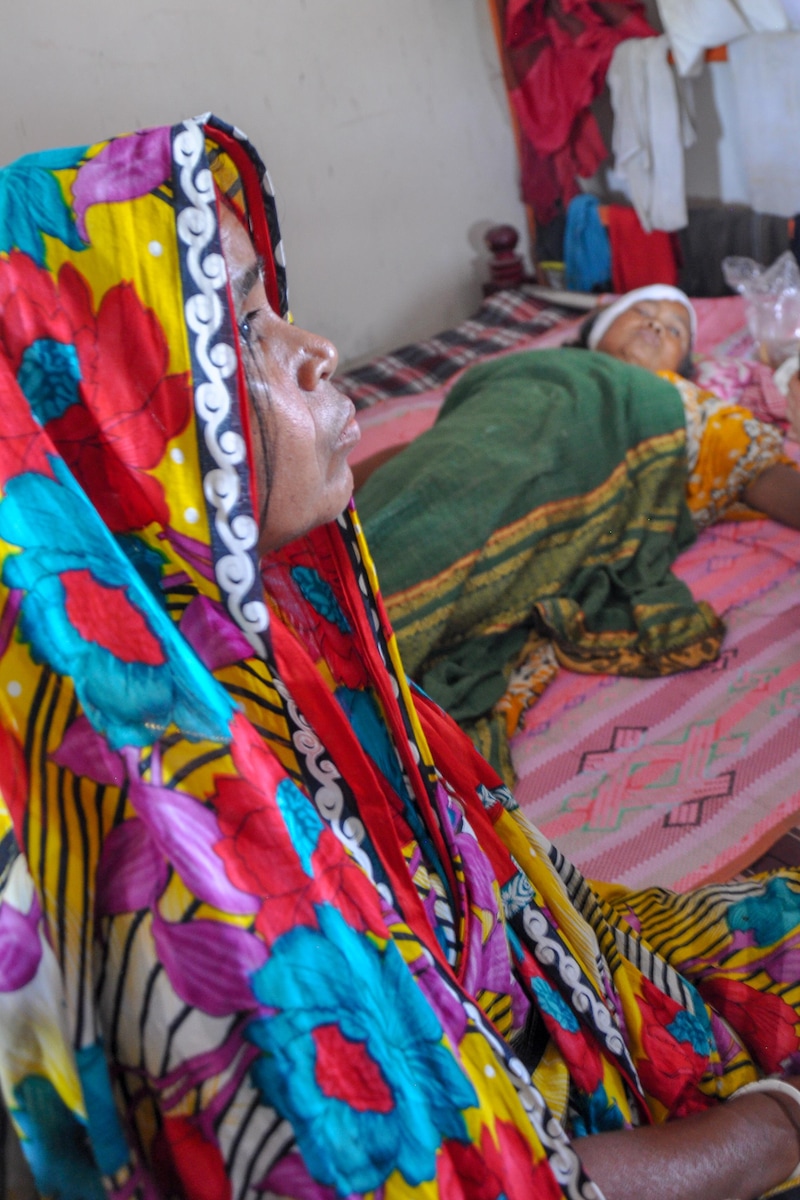 Une femme dans un lit la tête couverte d’un bandage ensanglanté. À côté, une femme a le regard triste.