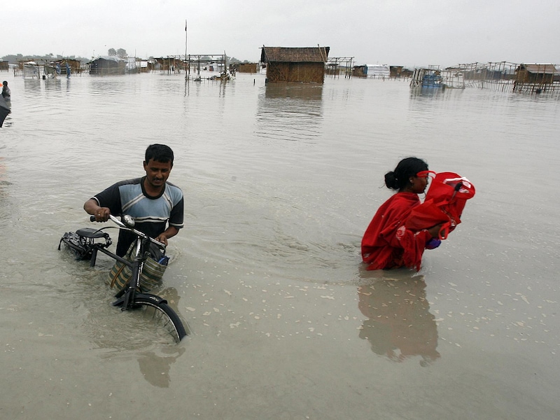 Une inondation détruit un village. Un homme pousse son vélo dans l’eau jusqu’à la taille. Une femme transporte ses biens.