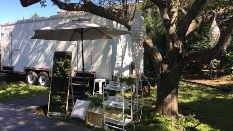 Sous un arbre, une roulotte blanche devant laquelle est disposé un parasol, un miroir et une petite étagère avec des produits. Sous un arbre, une roulotte blanche devant laquelle est disposé un parasol, un miroir et une petite étagère avec des produits.