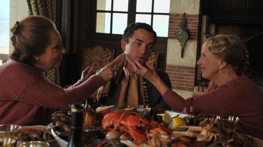 Autour d'une table, deux femmes habillées pareil, touchent le visage d'un jeune homme. Autour d'une table, deux femmes habillées pareil, touchent le visage d'un jeune homme.