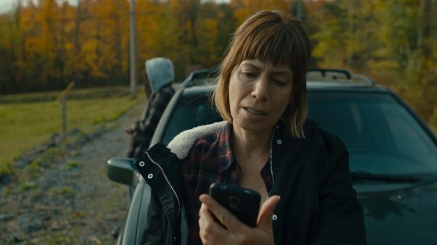 Une femme (Céline Bonnier) adossée contre une voiture regarde son téléphone.