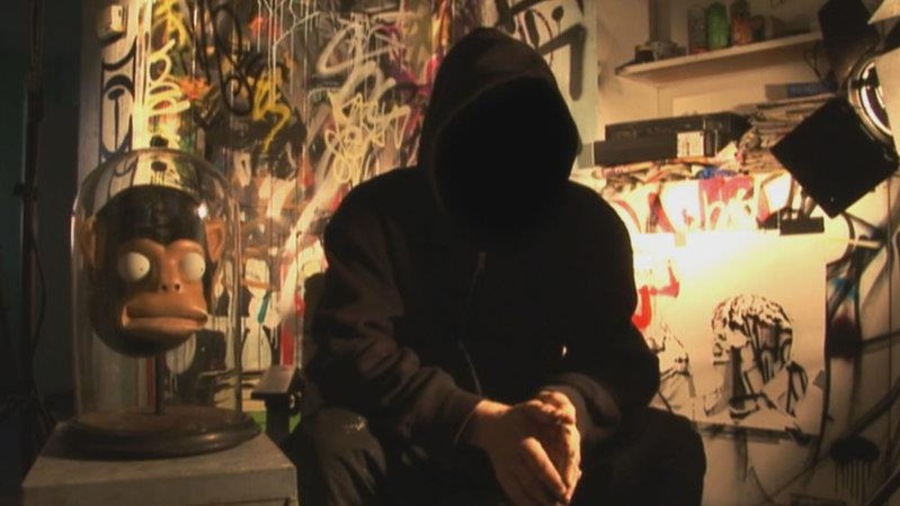 Un homme cagoulé, dont on ne voit pas le visage, assis dans un local plein de graffitis. Un homme cagoulé, dont on ne voit pas le visage, assis dans un local plein de graffitis.