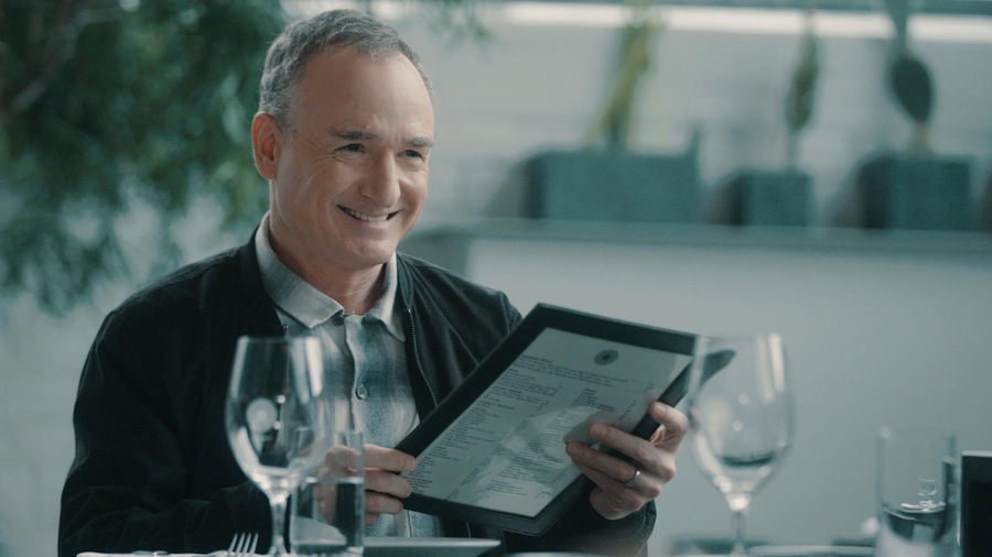 À table, Bruno Blanchet tient un menu dans ses mains et sourit.
