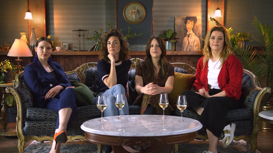 Quatre femmes assises dans un canapé antique, devant elles, une table contient quatre verres de vin blanc.