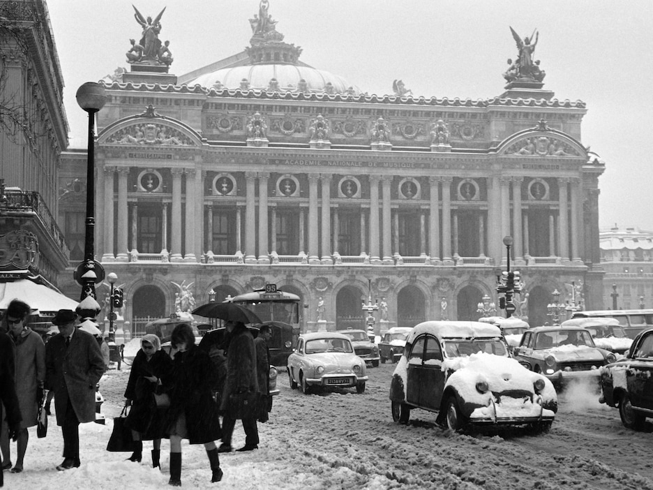 Photo en noir et blanc qui montre la place, des piétons et de nombreuses voiture empêtrées dans la neige. Photo en noir et blanc qui montre la place, des piétons et de nombreuses voiture empêtrées dans la neige.